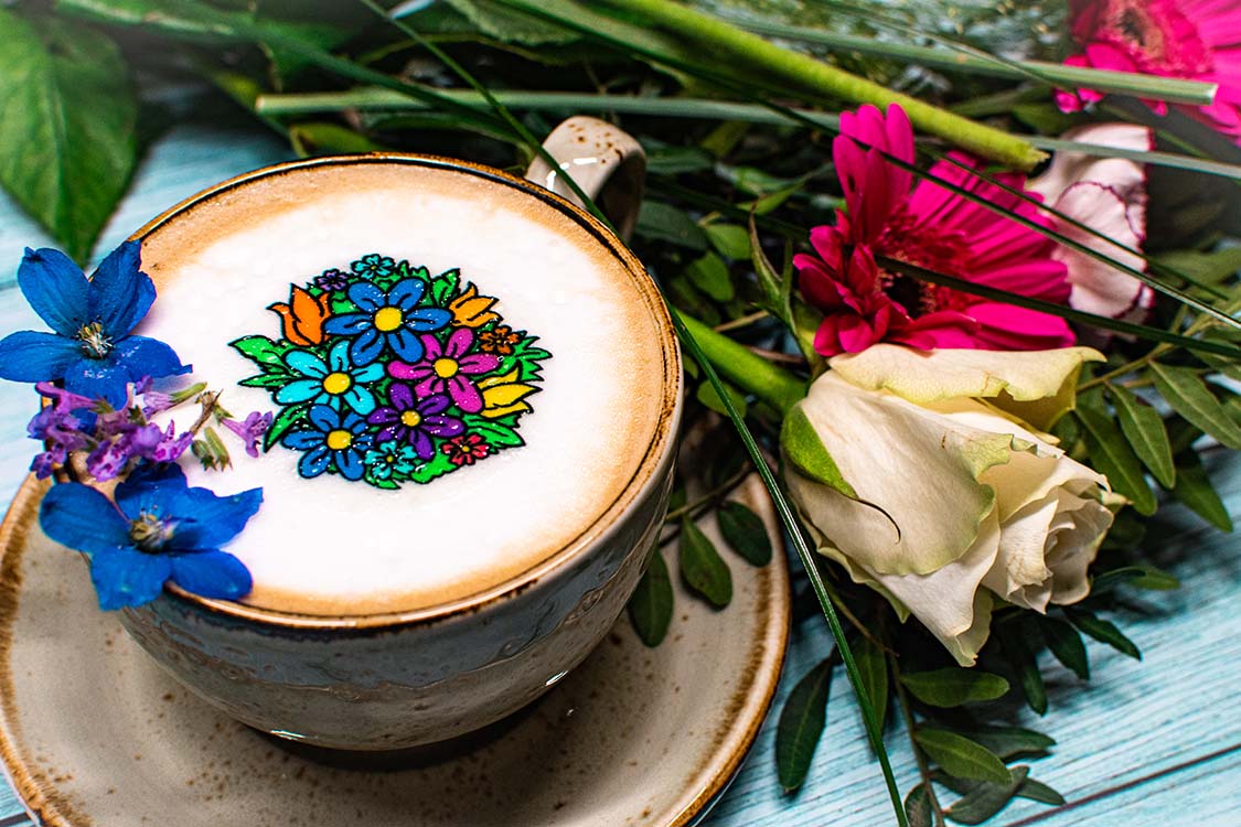 Blumenstrauß mit roten und weißen Rosen. Links steht eine graue Tasse mit passender Untertasse die mit einem Cappuccino gefüllt ist. Die Schaumkrone ist mit Blütenblätter und einem Blumenstrauß Twicky Sticky dekoriert.