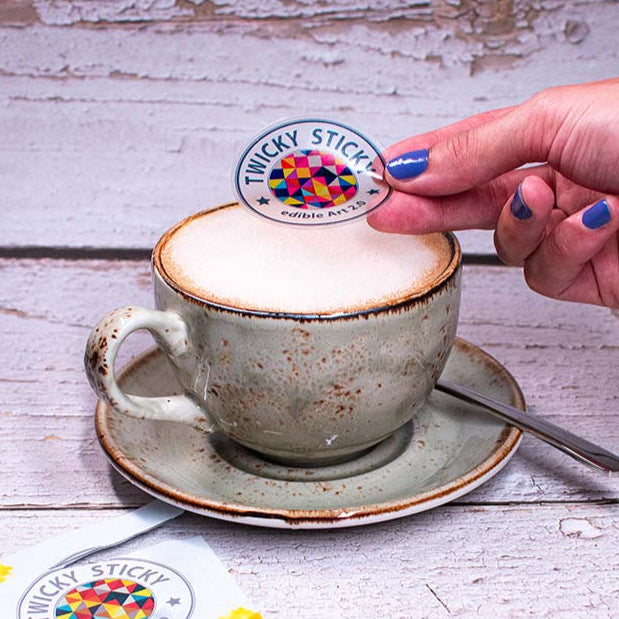Auf weißen Holzdielen steht die graue Kaffee Tasse. Eine Hand ist dabei den Milchschaum mit einem Twicky Sticky zu dekorieren