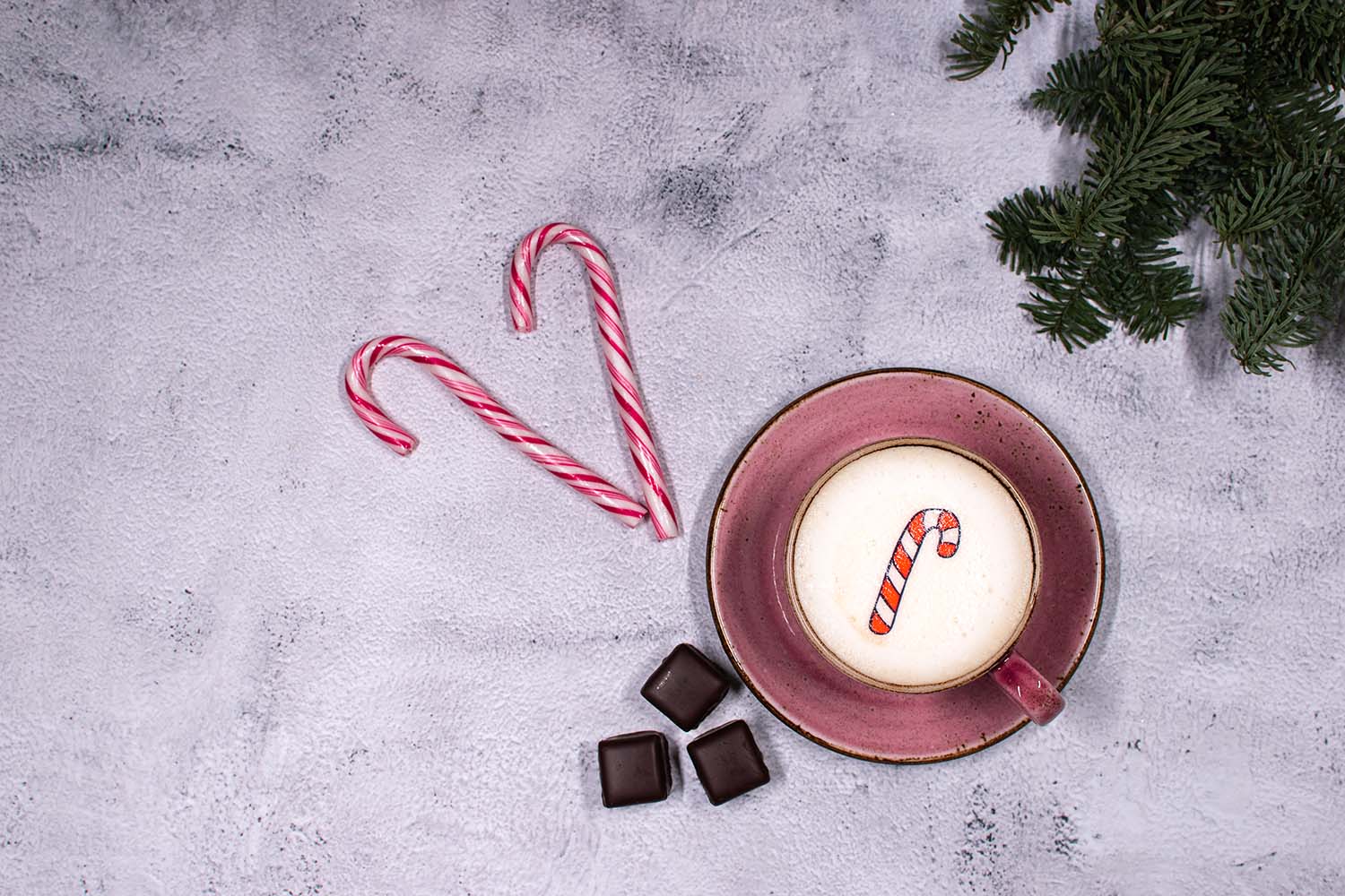Rote Tasse mitt Cappuccino und Zuckerstangen Twicky Sticky. Oben rechts im Bild sind Tannenzweige sowie Schokolade und Zuckerstangen 
