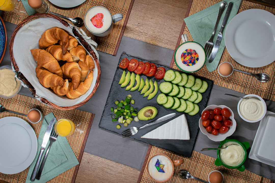 Zu sehen ist ein mit Crossaints, Gurken, Tomaten, Avocado, Kaese, Tomaten, Aufstrichen und Eiern gedeckter Fruehstueckstisch. Die Kaffeetassen sind dekoriert mit unterschiedlichen Motiven