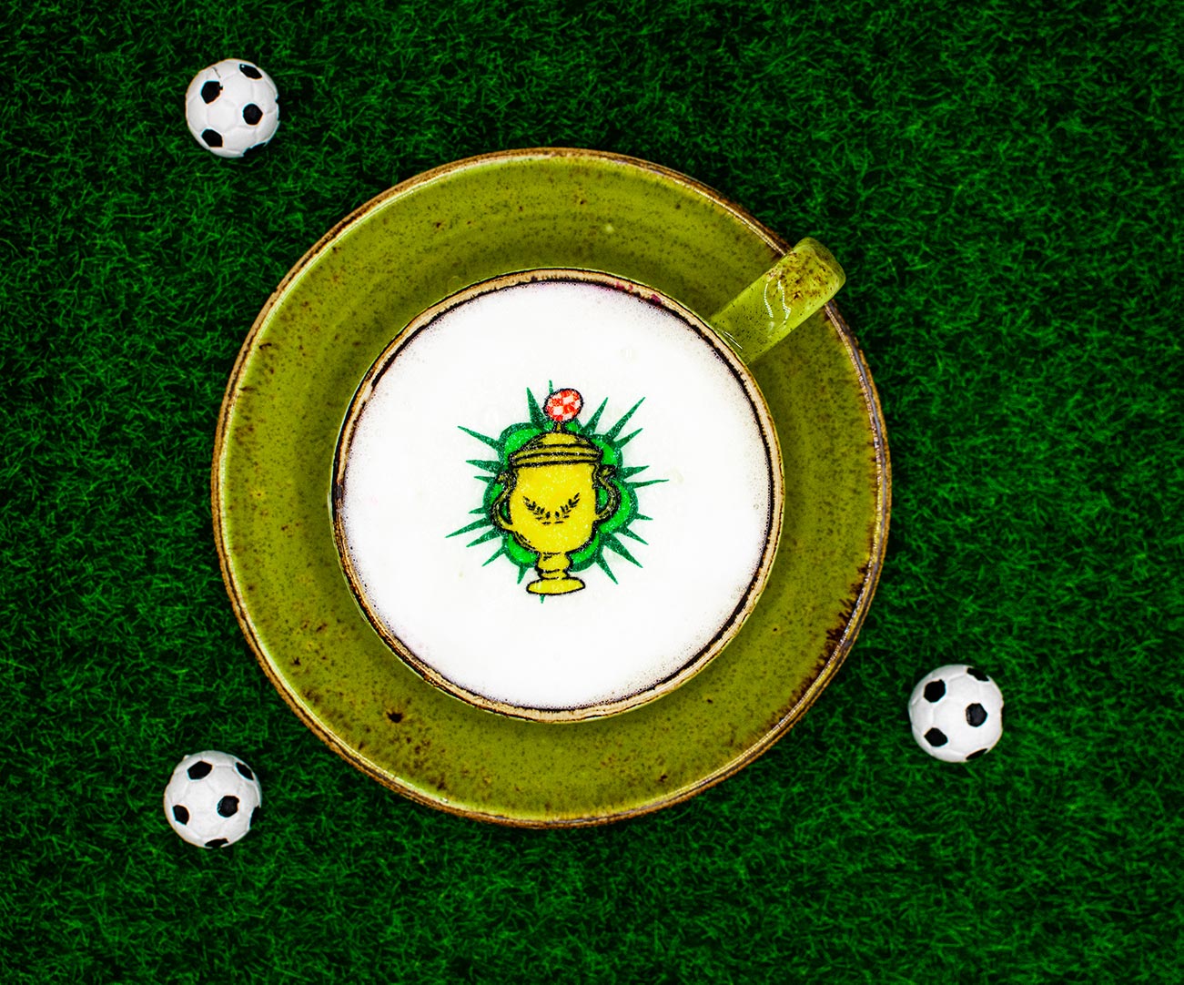 Der Hintergrund ist eine Wiese mit kleinen Fußbaellen. In der Mitte steht eine grüne Tasse mit Milchschaum, welcher mit einem Pokal dekoriert ist. 