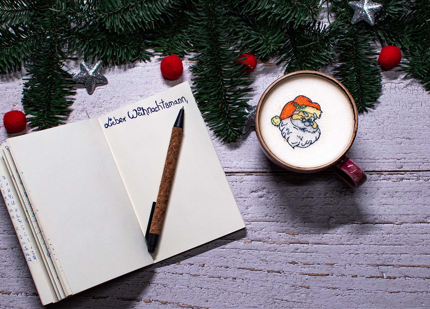 Notizbuch auf dem lieber Weihnachtsmann steht. Im Buch liegt ein Kugelschreiber und oben im Bild sind Tannenzweige mit Weihnachtskugeln. In einer roten Kaffeetasse ist Capuccino und Weihnachtsmann Sticker
