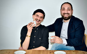 Die Gruender Peter Scherer und Burhan Yalcin sitzen mit Kaffee am Tisch und grinsen in die Kamera