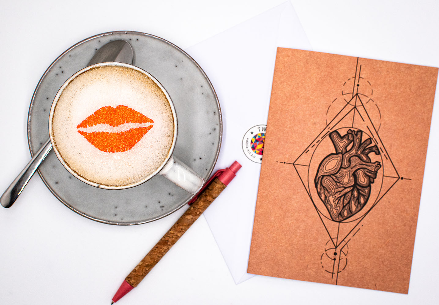 Twicky Sticky Kussmund auf Cappuccino in grauer Tasse. Rechts neben der Tasse Grußkarte anatomisches Herz mit Umschlag und Kugelschreiber