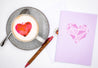 Twicky Sticky Watercolor Herz auf Cappuccino in grauer Tasse. Rechts neben der Tasse Grußkarte anatomisches Herz mit Umschlag und Kugelschreiber