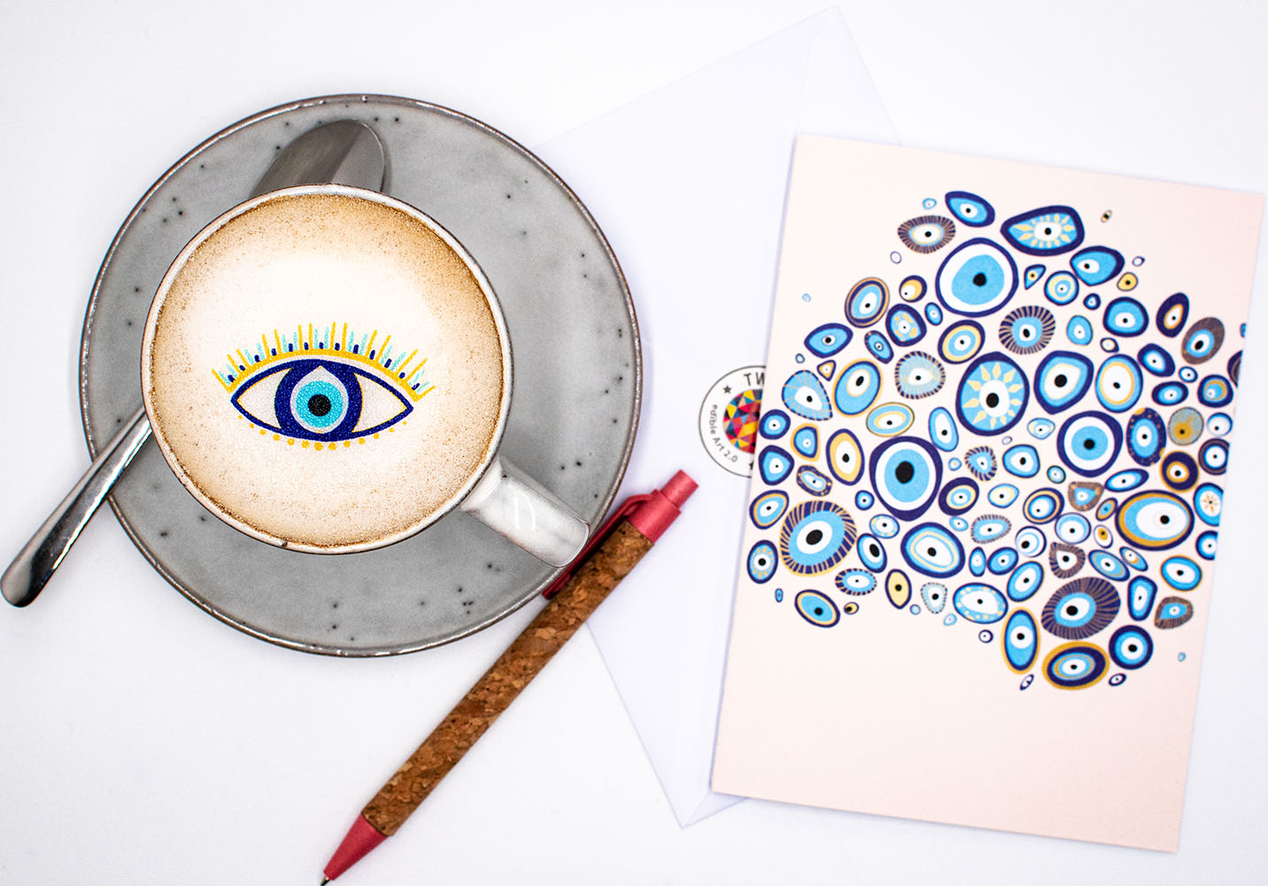 Twicky Sticky Nazar Auge auf Cappuccino in grauer Tasse. Rechts neben der Tasse Grußkarte anatomisches Herz mit Umschlag und Kugelschreiber