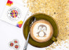 Twicky Sticky Pinguin in gruener Tasse mit Kaffee. Faltschachtel liegt neben der Untertasse und beige Steine sind rechts im Bild.